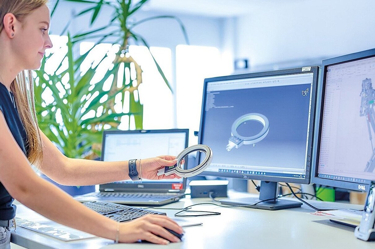 At the Factory: Female Mechanical Engineer Designs 3D Engine on Her Personal Computer. In the Background Male Automation Engineer who Uses Laptop for Programming Robotic Arm.