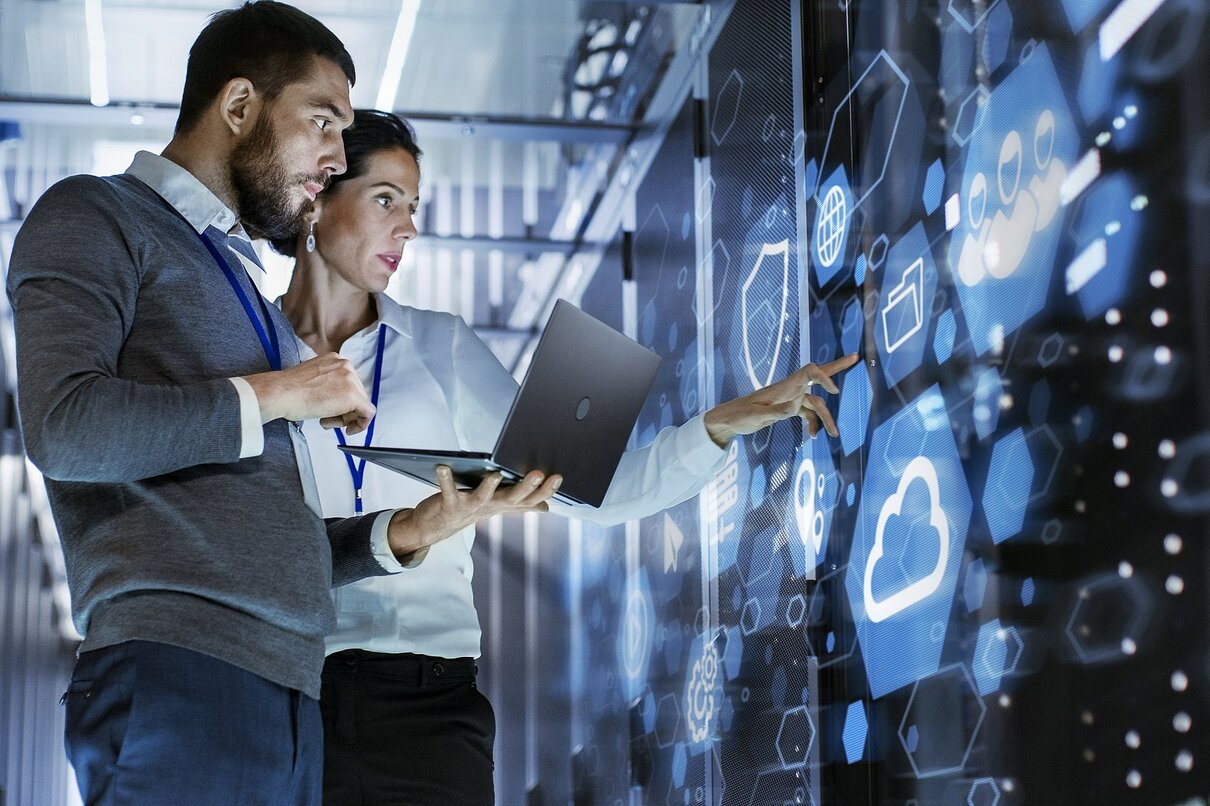 Male IT Specialist Holds Laptop and Discusses Work with Female Server Technician. They're Standing in Data Center, Rack Server Cabinet with Cloud Server Icon and Visualization.