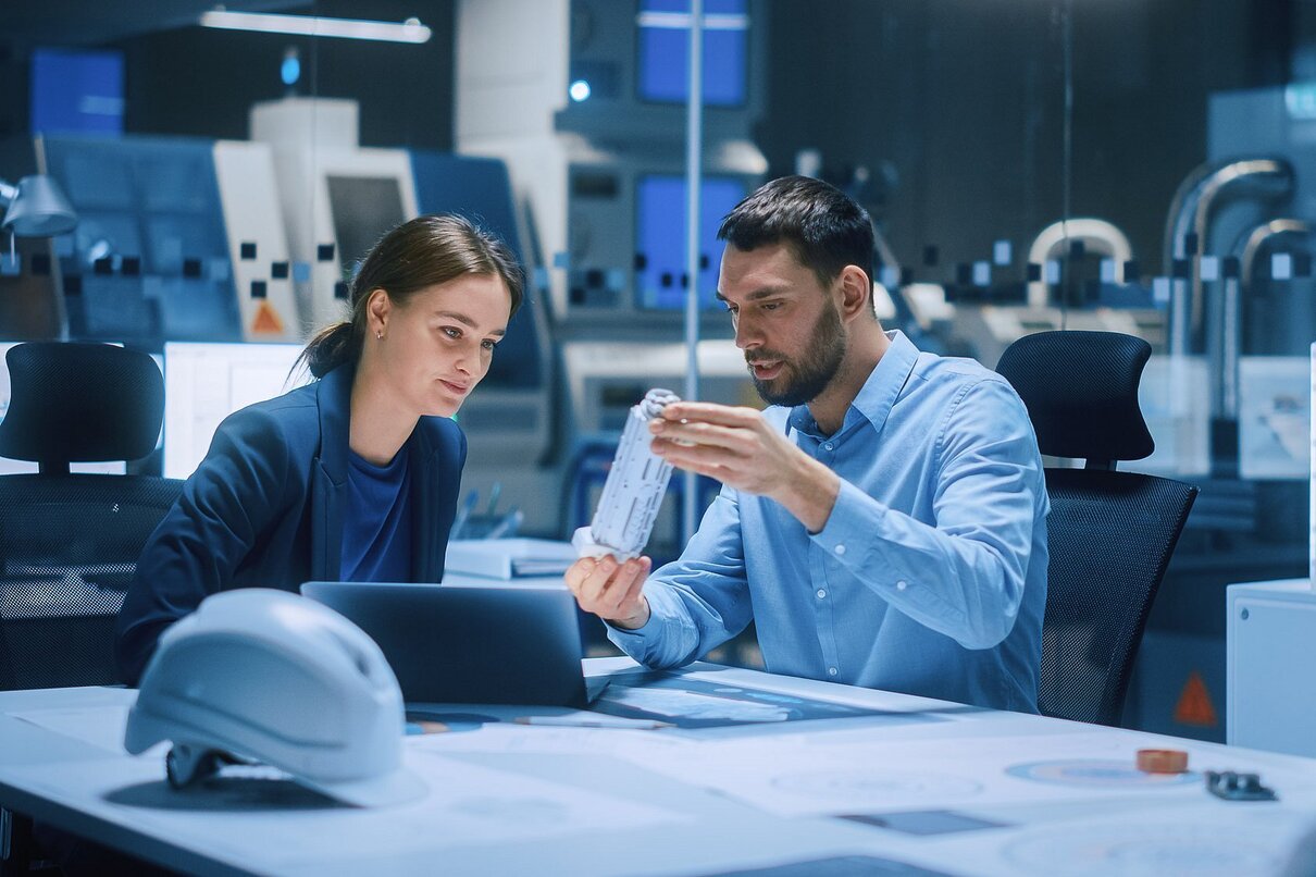 Industry 4.0 Modern Factory Meeting Room: Chief Engineer Holds Mechanism, Shows it to Female Designer, Use Laptop. Scientists in Contemporary Lab Build Electronic Machinery for With Futuristic Design